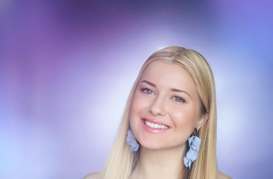 Smiling blonde woman adorned with delicate blue statement earrings, glowing against a dreamy purple and blue gradient background. Her elegance and warmth radiate through her relaxed expression and impeccable styling look