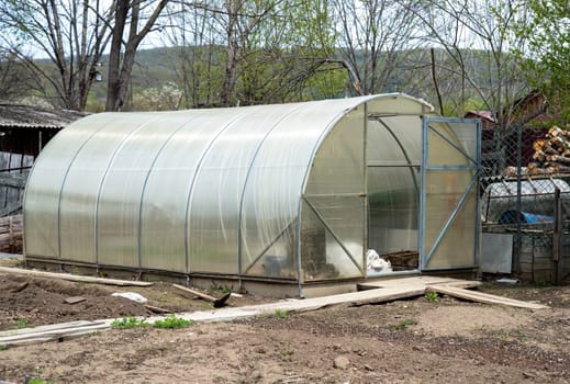 Open greenhouse in a backyard garden during spring. Transparent polycarbonate walls, surrounded by trees and gardening tools. Ideal for growing plants in rural settings