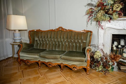 living room with beautiful antique sofa, fireplace and lamp. sofa with wooden legs, it is green in color, made of velvet fabric. on warm-colored parquet. around it is floral decor