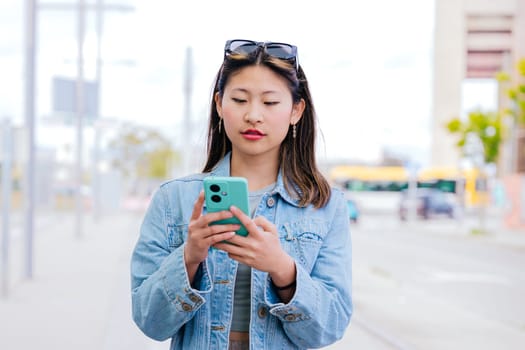 A stylish young woman in a denim jacket checks her smartphone while waiting at a tram station. She appears focused and relaxed, with sunglasses on her head. A modern urban lifestyle scene.
