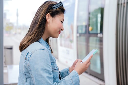 Side view of a young woman in a denim jacket using her smartphone while waiting to board a tram. Modern urban transportation and digital connectivity concept.