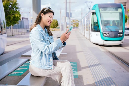 Cheerful young woman in a denim jacket sitting at a tram stop, using her smartphone while waiting for the approaching tram. Modern urban transportation and digital lifestyle concept.