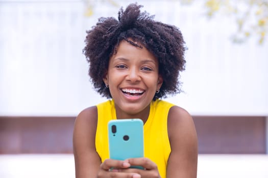 A young woman with natural curly hair wearing a yellow top rests her face on her hand while holding a smartphone. Calm and happy moment captured in a bright outdoor setting.