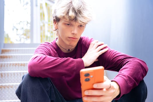 Young man in a burgundy shirt sitting on metal stairs, focused on his smartphone, with autumn leaves scattered around in an outdoor urban setting.
