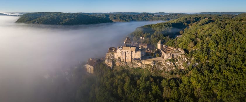 Chateau de Castelnaud sits atop a rocky outcrop in the Dordogne region, surrounded by morning fog as the sun begins to rise, creating a magical, serene atmosphere.