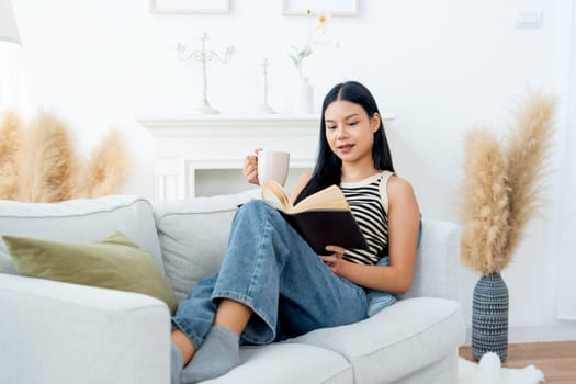 Front view beautiful Asian woman lean on sofa hold a cup of tea also enjoy to read the book with happiness in living room with day light.
