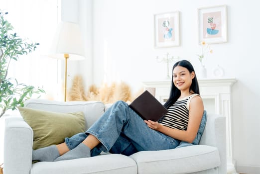 Side view and wide shot of beautiful Asian woman lean on sofa also enjoy to read the book and lood at camera with happiness on day light.