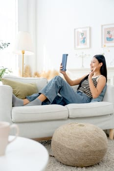 Vertical image of beautiful Asian woman lean on sofa also enjoy to use tablet in living room and express excited and happiness with day light.