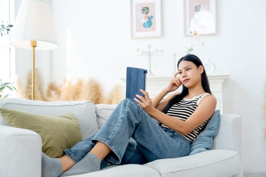 Wide shot of beautiful Asian woman lean on sofa also enjoy to use tablet in living room and express thinking action with day light.