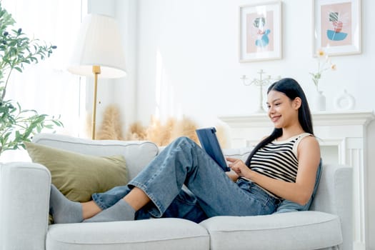 Side and high view beautiful Asian woman lean on sofa also enjoy to use tablet in living room with day light.