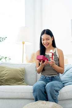 Vertical image of beautiful Asian woman sit on sofa in living room and hold box of present also look at the box and express happiness with  day light.