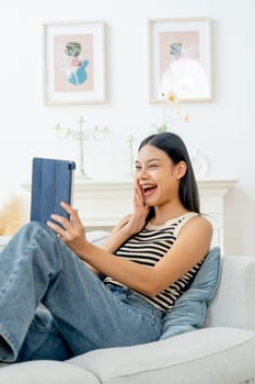 Vertical image of beautiful Asian woman lean on sofa also enjoy to use tablet in living room with day light.