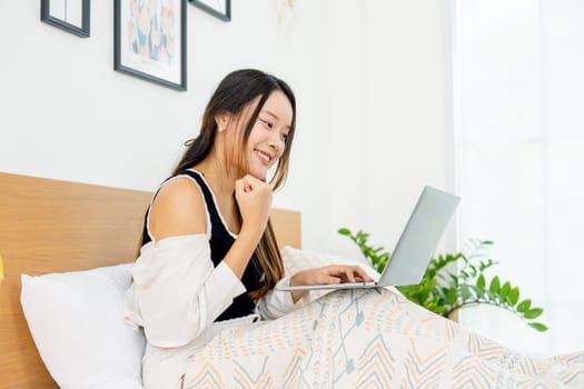 Side view of lovely Asian woman sit on bed and enjoy to use laptop for relaxing in bedroom.