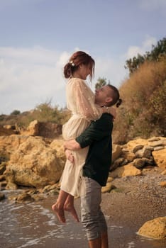 romantic and smiling couple guy holds girl in his arms, he lifted her up. very emotional photo. newlyweds happy and smiling. girl hugs guy by neck he lifted her up. around them sea and rocks