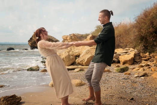 romantic and smiling couple dancing on the seashore. they are spinning barefoot on the seashore. the couple is holding hands, smiling and laughing. the sea and rocks are around them.