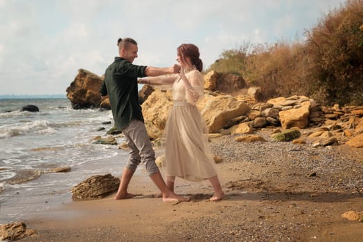 Romantic and smiling couple dancing on the seashore. They are barefoot, smiling and laughing. Around them are the sea and rocks. Girl in a delicate beige dress is dancing tango, horizontal orientation