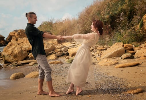 Romantic and smiling couple dancing on the seashore. They are spinning barefoot on the seashore. The couple is holding hands, smiling and laughing. Around them are sea waves and rocks.