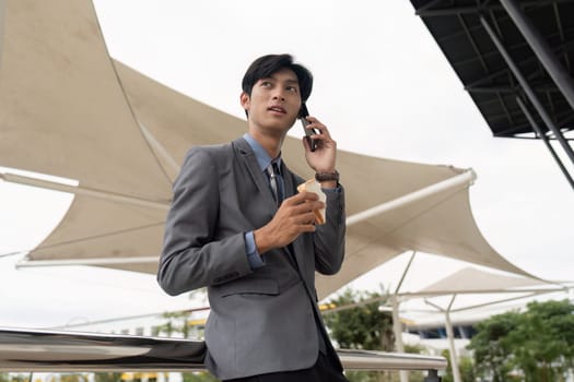 A young Asian businessman talks on his smartphone while enjoying ice cream outdoors, showcasing a blend of work-life balance and modern style in a contemporary setting.