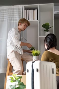 A young couple in their new home, one partner sitting on a stool, carefully placing plants on a shelf while the other watches, excited to decorate.