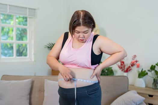 A plus size woman examines her waist measurement with a tape measure, thoughtfully considering her weight loss journey in a tidy living room setting.