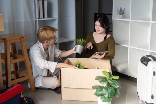 A young couple sits on the floor of their new living space, unpacking boxes and discussing where to place potted plants, filled with excitement.