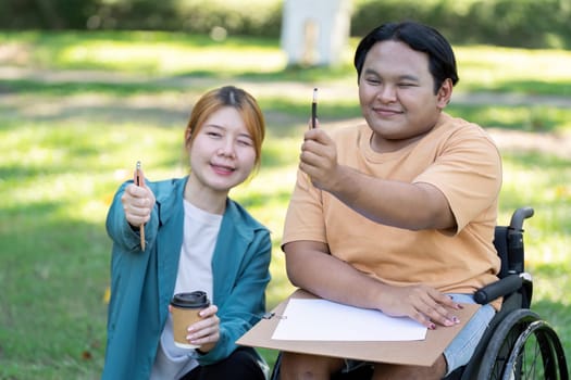 A smiling young man in a wheelchair holds up art tools alongside his supportive instructor, showcasing creativity and empowerment in a beautiful park setting.