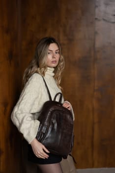 A girl in a warm sweater, a black short skirt, nylon tights, and black shoes is posing with a brown backpack