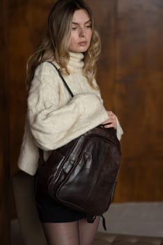 A girl in a warm sweater, a black short skirt, nylon tights, and black shoes is posing with a brown backpack