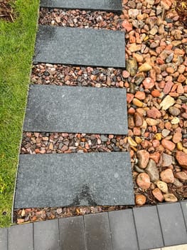 A stylish outdoor pathway with black rectangular stepping stones surrounded by colorful decorative rocks. This well-maintained landscaping design combines clean lines and natural elements, bordered by green grass and mulch with ornamental plants. A modern and functional addition to any outdoor space.
