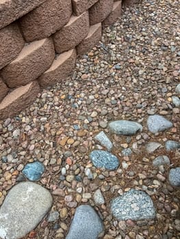 A close-up view of an aging retaining wall made of stacked concrete blocks, surrounded by a bed of decorative stones in various colors and sizes. The landscape shows signs of wear and is ready for renovation to restore its appeal and functionality.
