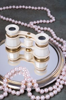Binoculars rest on a mirror surrounded by pearl necklaces. Elegance and timelessness.