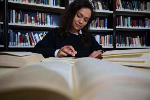 A focused young woman reads and studies among stacks of open books in a library.