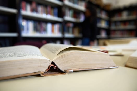 An open book rests on a library table, with shelves and a figure visible in the out-of-focus background. Represents knowledge, study, and the ambiance of reading spaces.