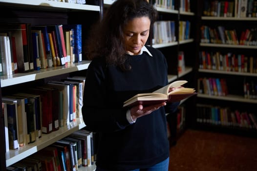 A person fully engrossed in reading a book while standing in a well-organized library, surrounded by bookshelves filled with a variety of colorful books.