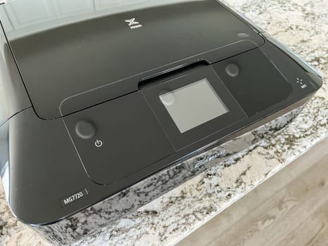 Castle Rock, Colorado, USA-May 8, 2023-A close-up image of a Canon printer placed on a granite countertop. The printer black glossy exterior prominently displays the Canon logo, reflecting light. The countertop beneath showcases a modern kitchen setting, blending functionality with sleek design.