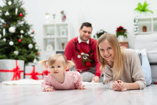 In the foreground a woman and a little girl lying on the floor. In the background is a blurry figure of a man wearing a Christmas sweater with an image of a reindeer. There is a dressed Christmas tree in the room and presents under it.