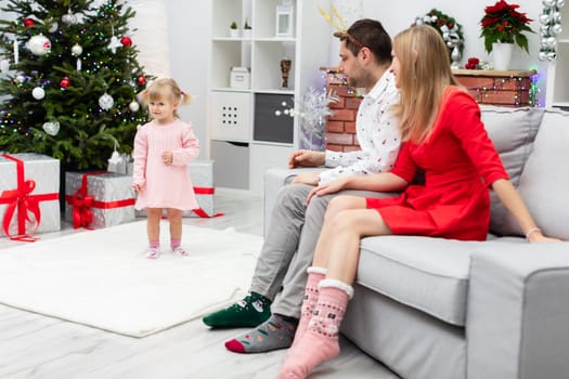 A mom, dad and a little daughter are together in a festively decorated room. The little girl stands on a fluffy white carpet. The parents are sitting on a gray couch facing the Christmas tree.