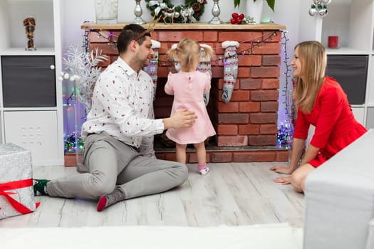 A man and a woman are sitting on the floor by a brick fireplace. Between them stands a little girl. The girl is facing away from the fireplace. The whole family is wearing festive clothes