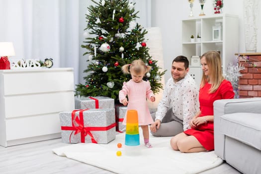 Mom and dad are sitting on the carpet, not far from the Christmas tree. Next to her parents stands a little girl. The child is wearing a pink dress. The parents are playing with the child in stacking a tower.