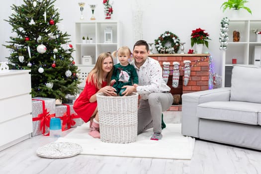 Parents with their little daughter are staying in a decorated room. The room has lots of Christmas decorations. The parents are smiling and the little girl is sitting in a wicker basket.