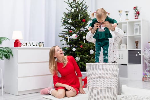 A dad lifts up his little daughter. A man takes the baby out of a wicker basket. A woman in a red dress sits on the floor and laughs with her mouth open. The room is decorated with Christmas ornaments.