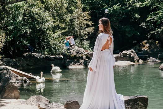 a beautiful woman in a long white dress looks into the distance at a beautiful lake with swans