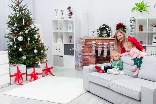 A woman in a red dress stands behind a gray couch and smiles. Sitting on the couch are two small children: a boy and a girl. In the background you can see a fireplace decorated with lights and a decorated Christmas tree with presents.