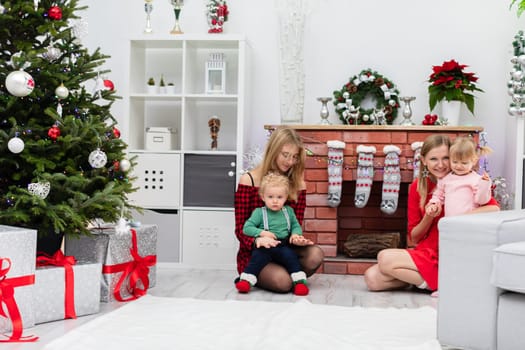The brick fireplace is profusely decorated with Christmas decorations. Wood lies in the fireplace. A woman sits by the fireplace and hugs a little girl in a pink dress. Next to her sits another woman, who holds a little boy on her lap.