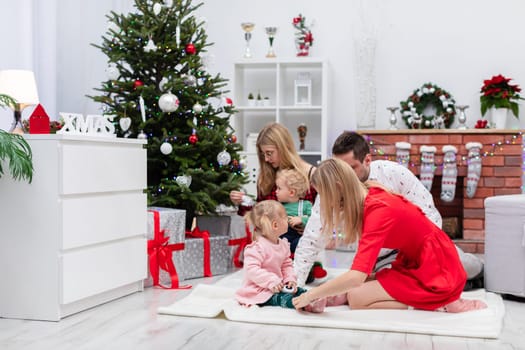 Two women, a man and two young children spend Christmas together. The family is playing on the carpet by the Christmas tree. The room is decorated with Christmas ornaments. Under the Christmas tree lie gifts.