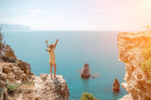 Happy girl stands on a rock high above the sea, wearing a yellow jumpsuit and sporting braided hair, depicting the idea of a summer vacation by the sea