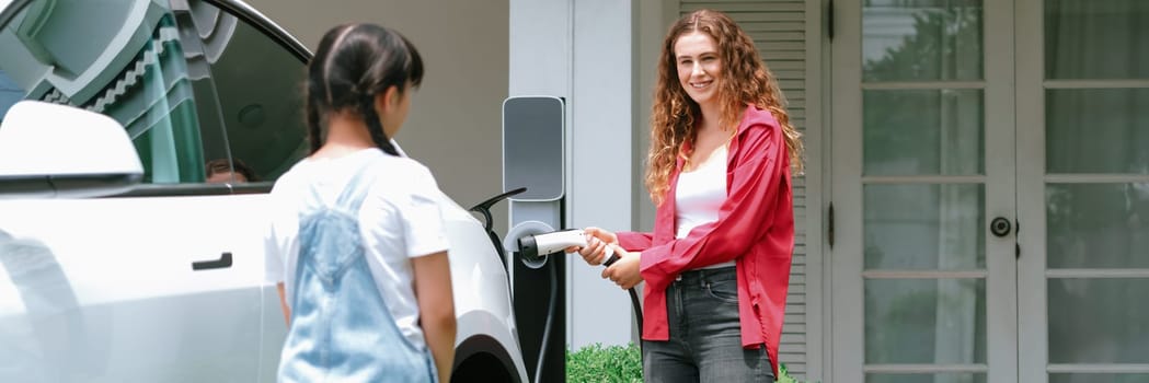 Happy little young girl learn about eco-friendly and energy sustainability as she help her mother recharge electric vehicle from home EV charging station. EV car and modern family. Panorama Synchronos