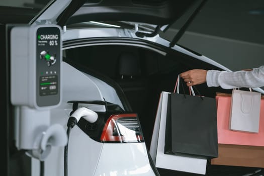 Young woman travel with EV electric car to shopping center parking lot charging in downtown city showing urban sustainability lifestyle by green clean rechargeable energy of electric vehicle innards