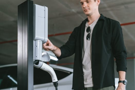 Young man travel with EV electric car to shopping center parking lot charging in downtown city showing urban sustainability lifestyle by green clean rechargeable energy of electric vehicle innards