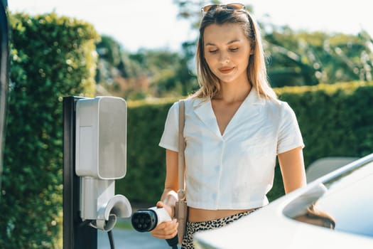 Young woman travel with EV electric car charging in green sustainable city outdoor garden in summer. Urban sustainability lifestyle by green clean rechargeable energy of electric BEV vehicle innards